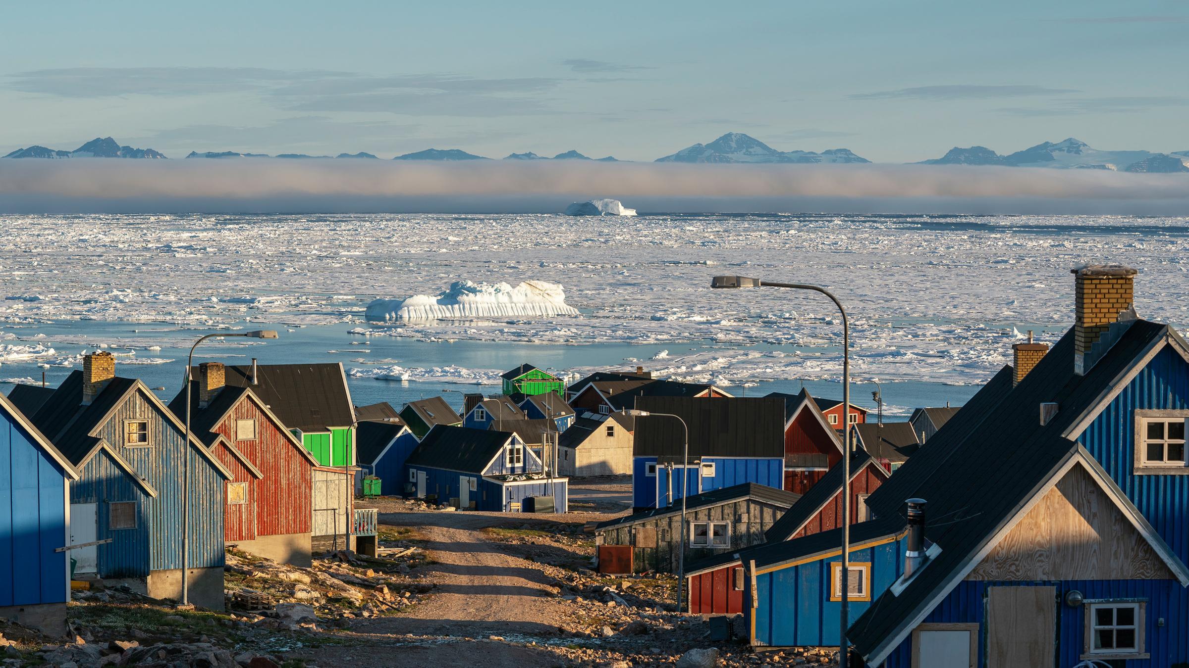 Bunte Holzhäuser getrennt druch eine Schotterstraße, im Hintergrund Eismeer mit von Nebel bedeckten Bergen.