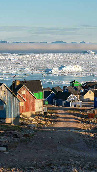 Bunte Holzhäuser getrennt druch eine Schotterstraße, im Hintergrund Eismeer mit von Nebel bedeckten Bergen.