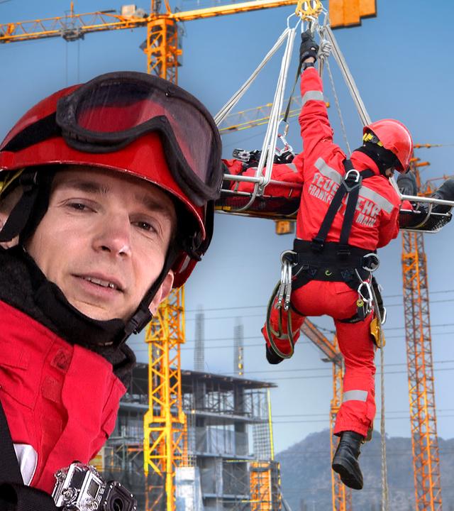 Eric mit Helm und Schutzkleidung, hinter ihm seilt sich ein Feuerwehrmann an einem Kran ab.