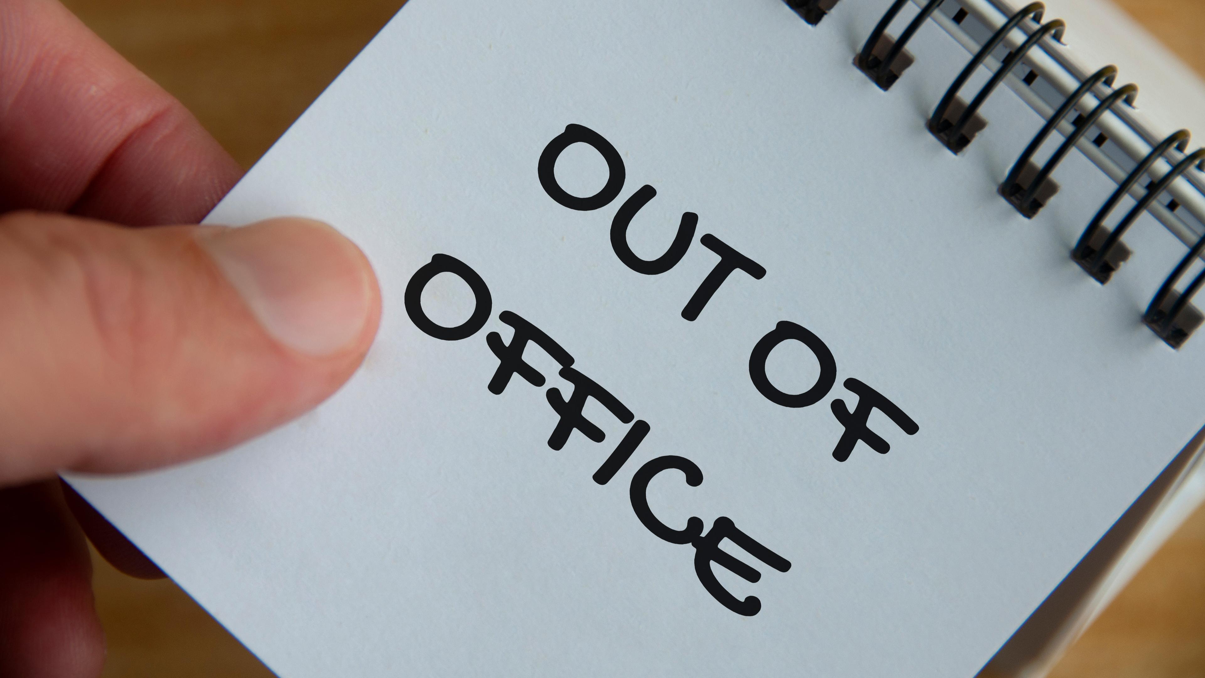 Eine Hand hält einen Notizblock, auf dem "Out of Office" geschrieben steht.