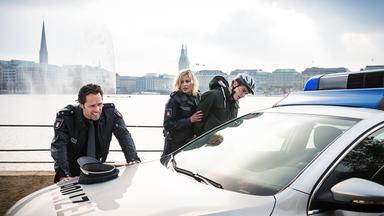 Der Polizeibeamte Mattes Seeler (Matthias Schloo) stützt sich mit beiden Händen auf der Motorhaube des Polizeiwagens ab. Die Polizeibeamtin Melanie Hansen (Sanna Englund) verhaftet Nils Kosta (Marin Bruchmann)