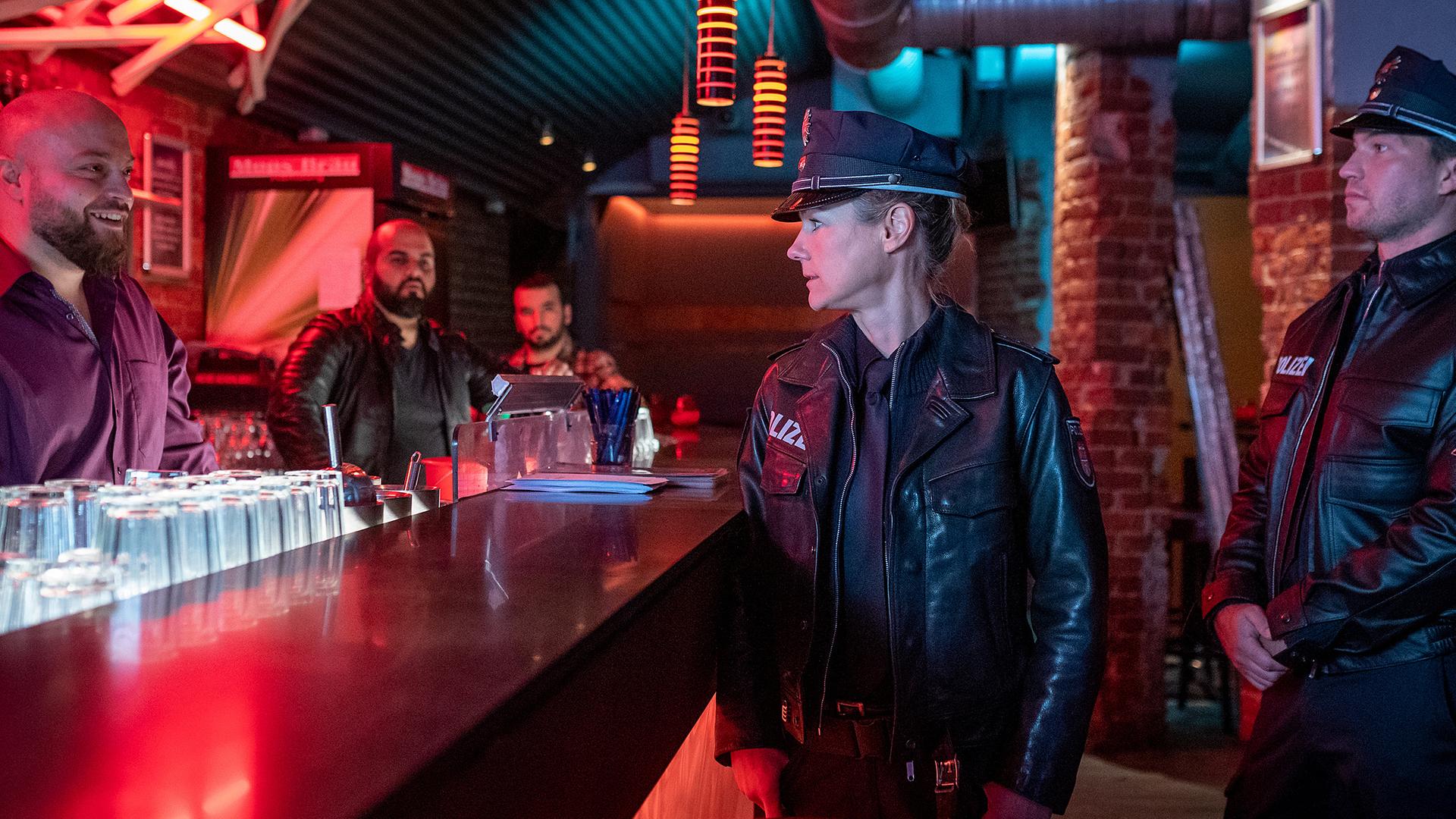 Die Polizistin Franzi Jung (Rhea Harder-Vennewald) und ihr Kollege Nick Brandt (Raúl Richter) stehen in einer Bar vor der Theke. Sie sehen zu Milo Novak (Baris Ar), der grinsend hinter der Theke steht. Im Hintergrund stehen zwei weitere Männer (Komparsen).