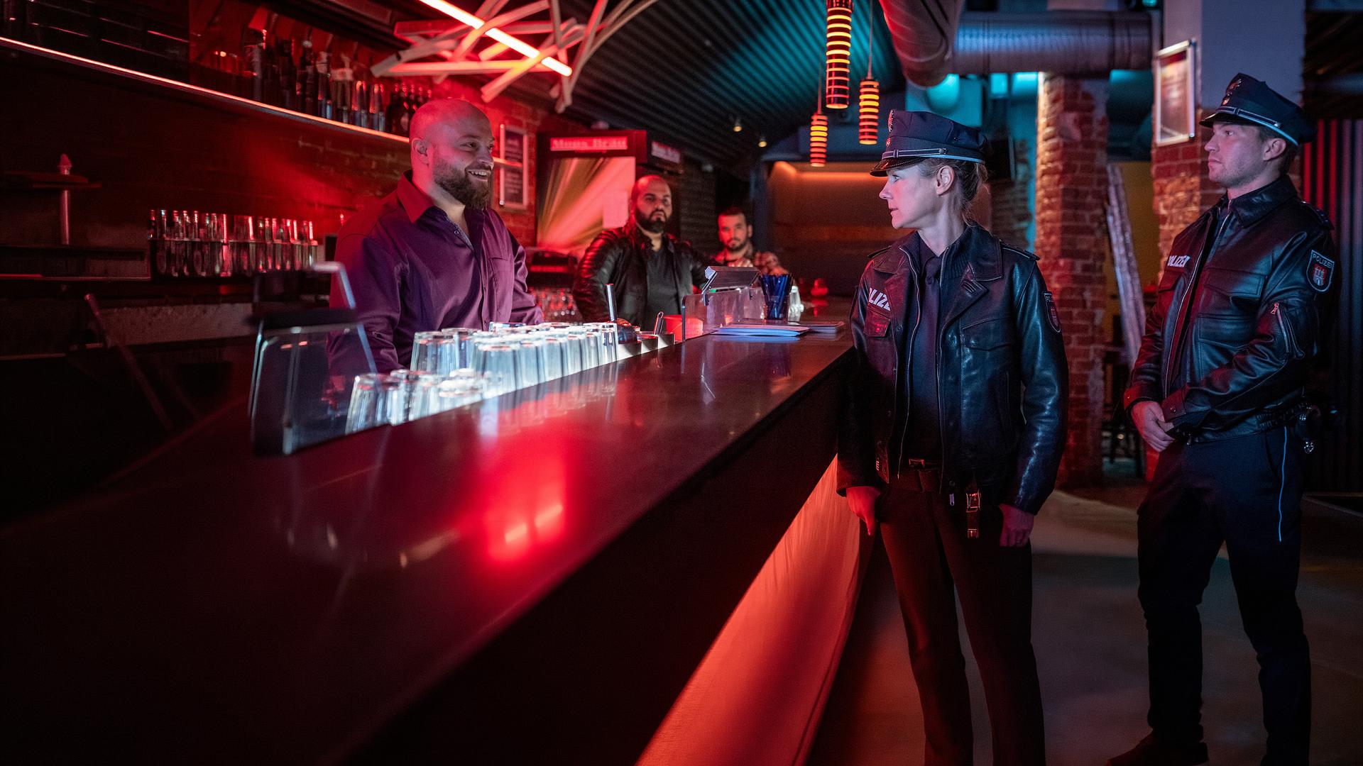 Die Polizistin Franzi Jung (Rhea Harder-Vennewald) und ihr Kollege Nick Brandt (Raúl Richter) stehen in einer Bar vor der Theke. Sie sehen zu Milo Novak (Baris Ar), der grinsend hinter der Theke steht. Im Hintergrund stehen zwei weitere Männer (Komparsen).
