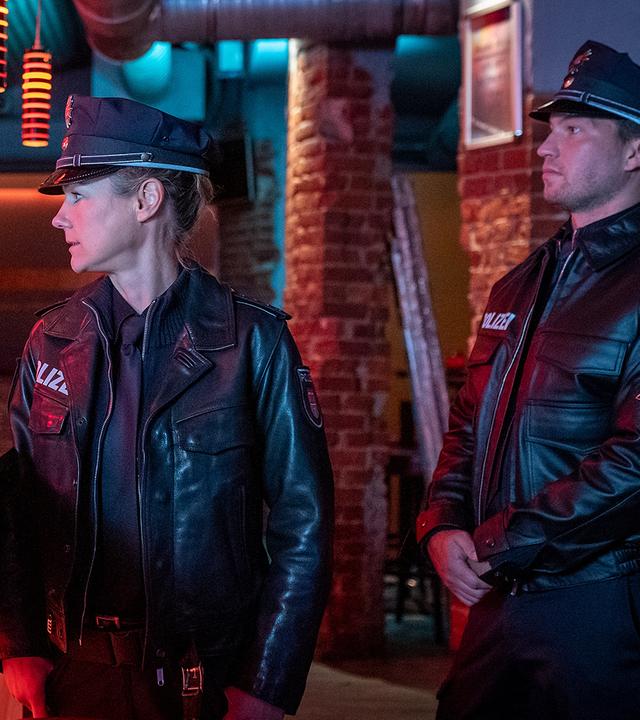 Die Polizistin Franzi Jung (Rhea Harder-Vennewald) und ihr Kollege Nick Brandt (Raúl Richter) stehen in einer Bar vor der Theke. Sie sehen zu Milo Novak (Baris Ar), der grinsend hinter der Theke steht. Im Hintergrund stehen zwei weitere Männer (Komparsen).