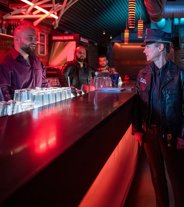 Die Polizistin Franzi Jung (Rhea Harder-Vennewald) und ihr Kollege Nick Brandt (Raúl Richter) stehen in einer Bar vor der Theke. Sie sehen zu Milo Novak (Baris Ar), der grinsend hinter der Theke steht. Im Hintergrund stehen zwei weitere Männer (Komparsen).