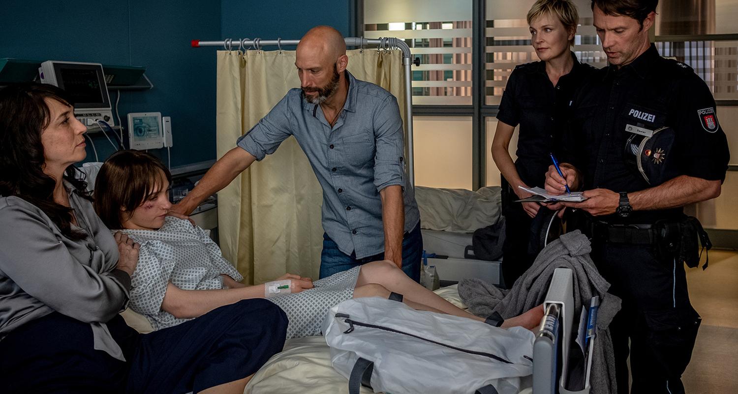 "Notruf Hafenkante - Ich heiße Maja": Im Krankenzimmer liegt Maja (Michelangelo Fortuzzi) in ihrem Bett. Auf ihrer linken Seite sitzt ihre Mutter Susanne (Jele Brückner). Auf der rechten Seite des Bettes stehen ihr Vater Torben (Andreas Grötzinger) und die Polizisten Melanie Hansen (Sanna Englund) und Mattes Seeler (Matthias Schloo). Mattes schreibt etwas mit einem Kugelschreiber in sein Notizbuch.