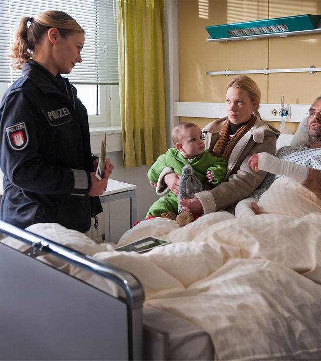 "Notruf Hafenkante - Leben daneben": Franzi (Rhea Harder) befragt Annik Karberg (Theresa Hübchen) und ihren Freund Enno Rüfken (Jophi Ries), der im Krankenbett liegt. Annik hat ein Kleinkind auf dem Schoß.
