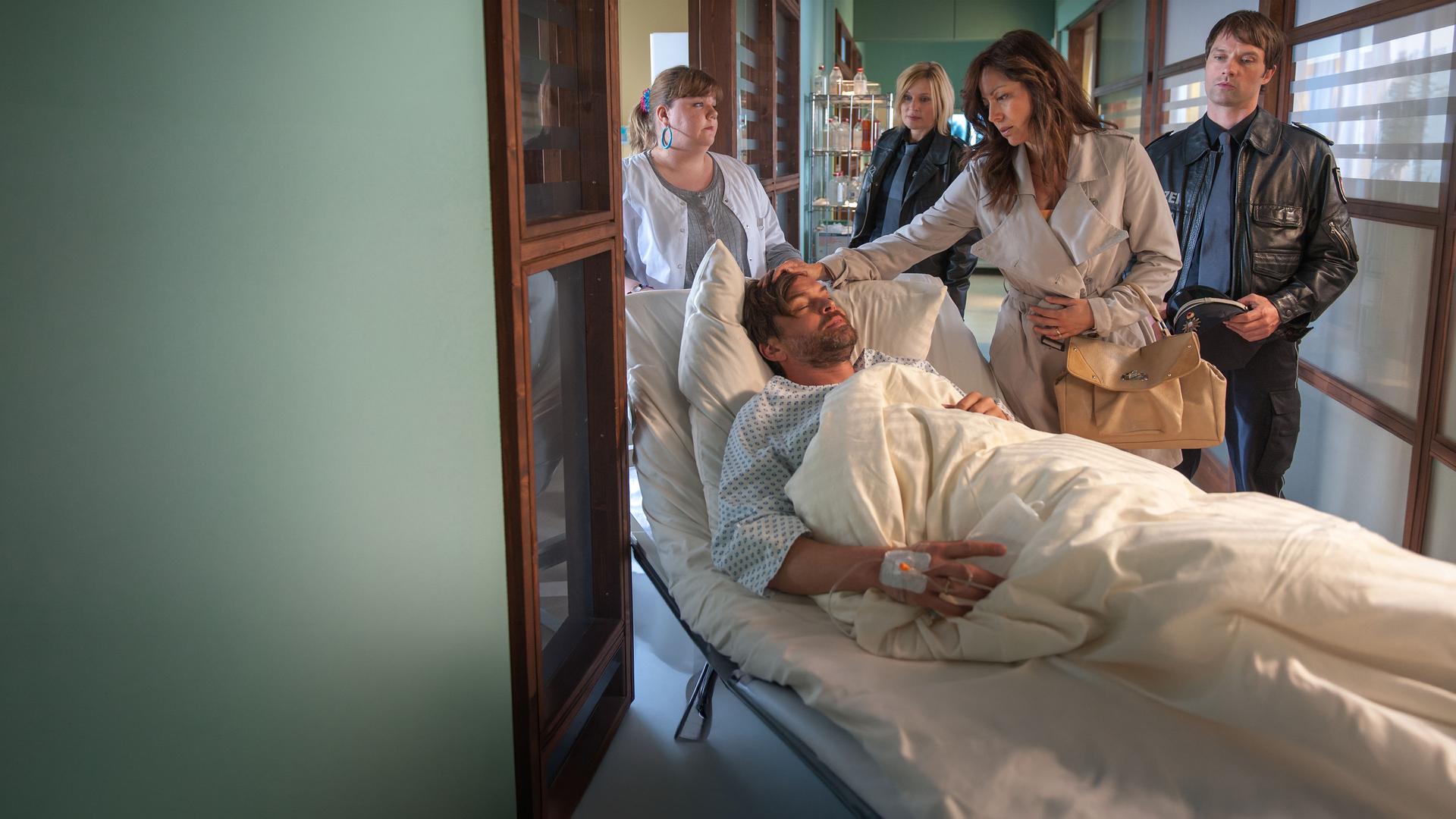 "Notruf Hafenkante - Lügen und Geheimnisse": Frauke (Manuela Wisbeck) schiebt Torsten Leitner (Ole Puppe) in einem Krankenbett über einen Flur des EKH. Neben dem Bett steht Miriam Leitner (Carolina Vera), die ihrem Mann besorgt über den Kopf streicht. Hinter ihr stehen Melanie (Sanna Englund) und Mattes (Matthias Schloo).