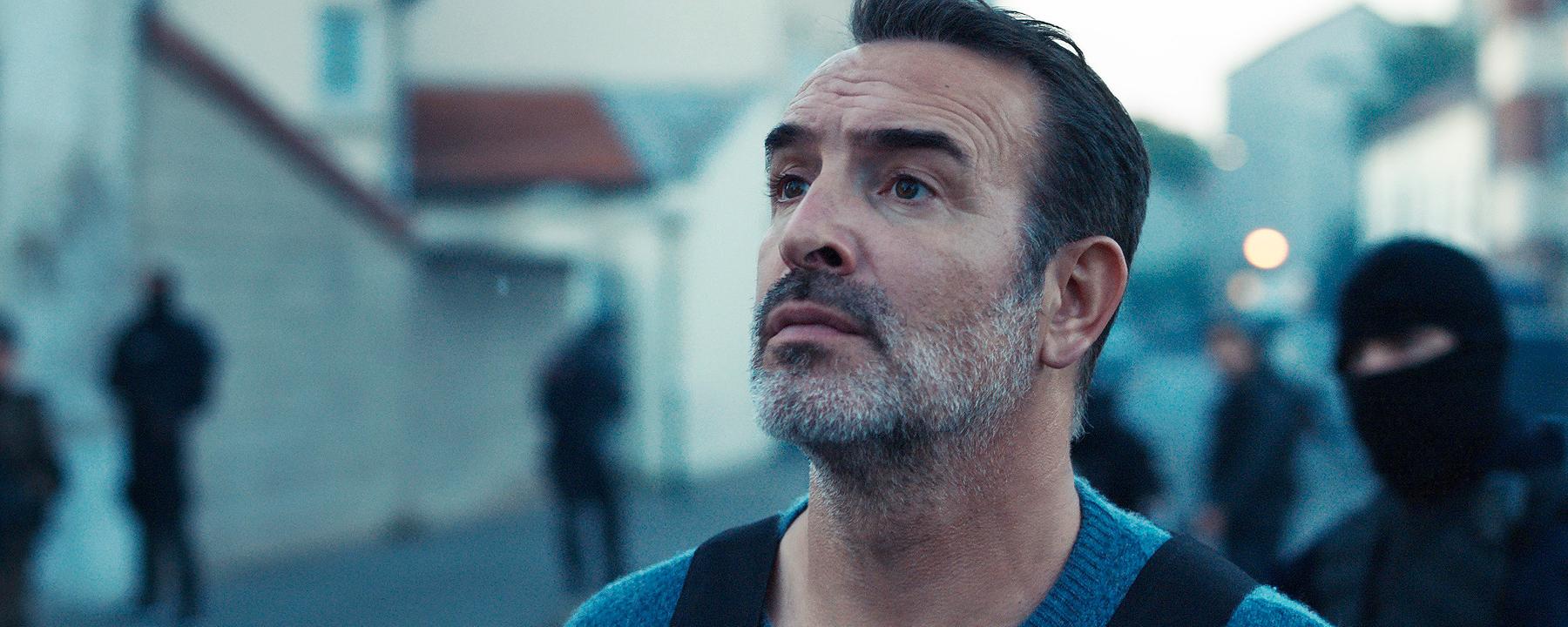 Fred (Jean Dujardin) steht in einer schusssicheren Weste im Morgengrauen auf einer Straße, hinter ihm stehen einige vermummte Männer einer Spezialeinheit.