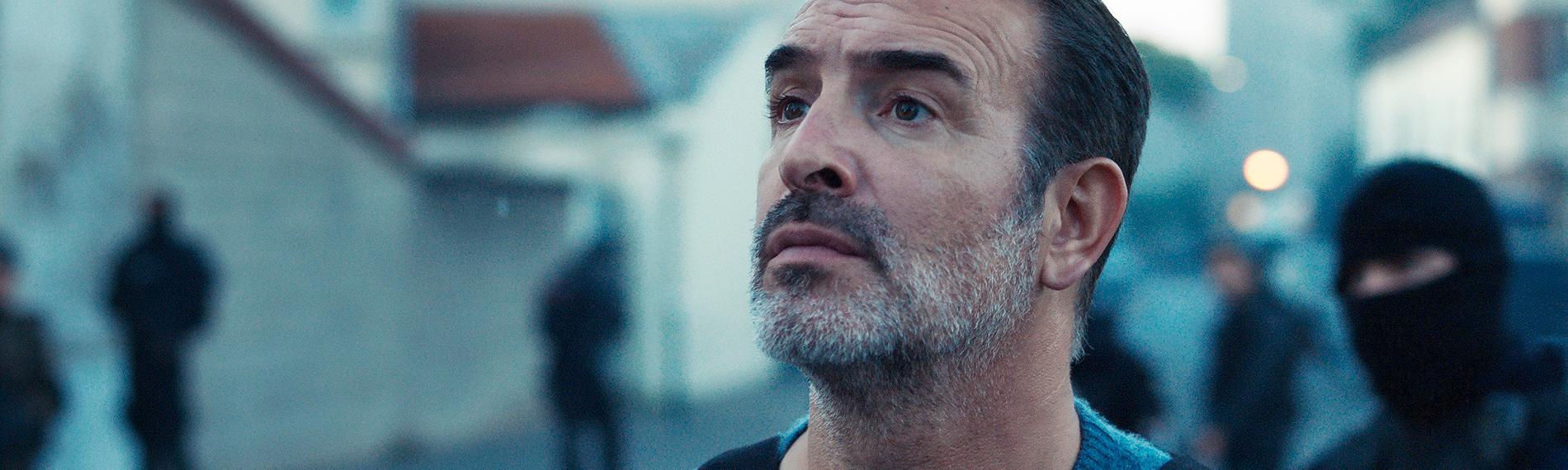 Fred (Jean Dujardin) steht in einer schusssicheren Weste im Morgengrauen auf einer Straße, hinter ihm stehen einige vermummte Männer einer Spezialeinheit.