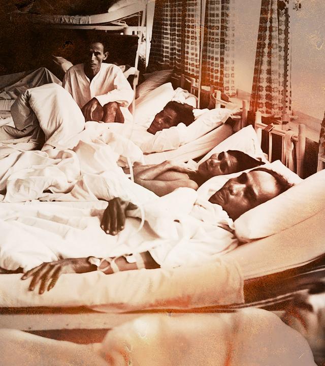 Mehrere Männer liegen in einem engen Raum in Betten nebeneinander, zugedeckt mit weißen Laken; einige schauen zur Kamera, einer sitzt aufrecht  Szene aus einem Lazarettschiff.