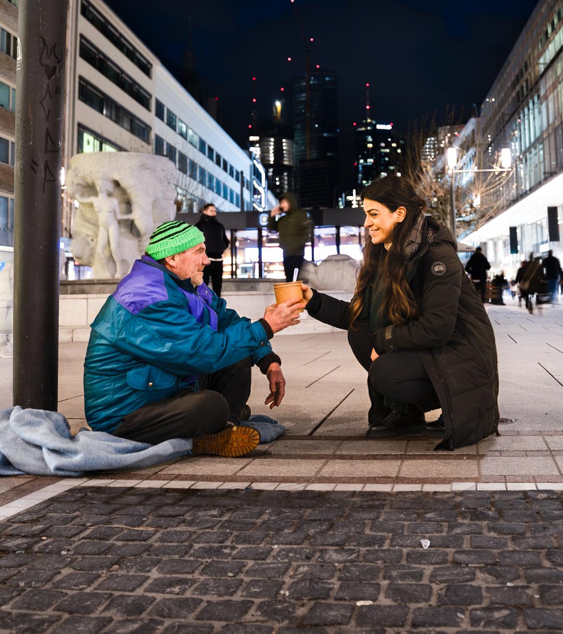 Es ist Nacht in der Frankfurter Fußgängerzone. Im Vordergrund sitzt ein obdachloser Mann mit grüner Mütze. Vor ihm kniet eine junge Frau mit langen schwarzen Haaren, die dem Obdachlosen einen Mahlzeit überreicht.