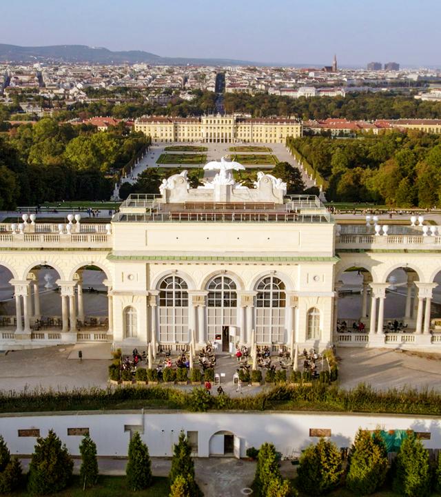 Das Bild zeigt die Gloriette, ein architektonisches Bauwerk im Schönbrunner Schlosspark in Wien, Österreich. Die Gloriette ist auf einem erhöhten Platz errichtet und bietet einen weiten Blick über die Stadt Wien und die umliegenden Landschaften.   Im Vordergrund sind die beeindruckenden Säulen und Bögen der Gloriette zu sehen, die in einem hellen Farbton gehalten sind. Die Fassade ist reich verziert, und über dem Hauptteil des Gebäudes befinden sich Skulpturen, darunter figürliche Darstellungen.  Unterhalb der Gloriette sind Besucher zu erkennen, die auf einer Terrasse sitzen und die Aussicht genießen. Im Hintergrund zieht sich die Stadt Wien mit ihren charakteristischen Gebäuden und Bäumen in die Ferne. Der Himmel ist klar, was eine helle und freundliche Atmosphäre schafft.   Die gesamte Szene vermittelt ein Gefühl von historischer Eleganz und natürlicher Schönheit, umgeben von der Pracht der österreichischen Landschaft.