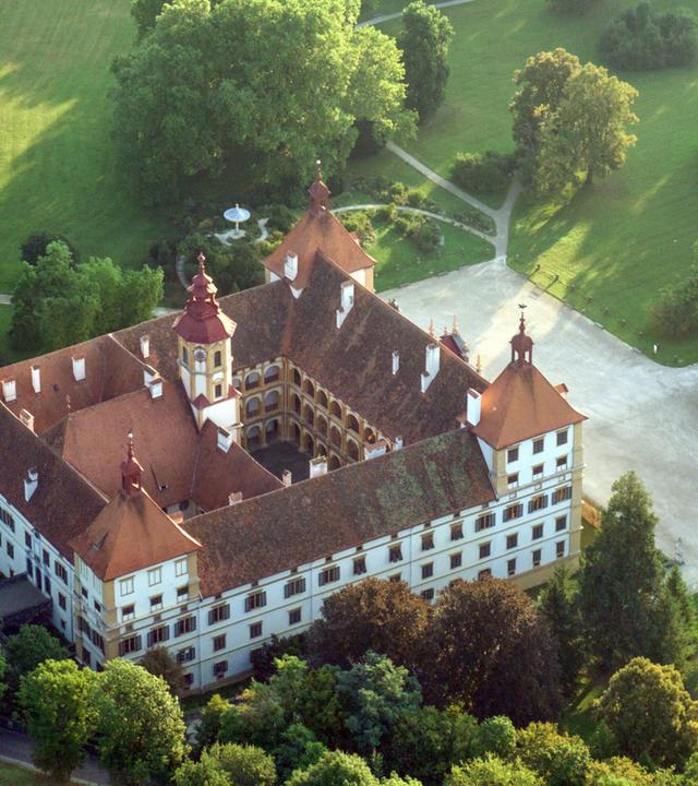Das Bild zeigt das Schloss Eggenberg in Graz, Österreich, aus einer luftigen Perspektive. Das Schloss ist von großen, grünen Parkanlagen umgeben, die mit vielen Bäumen und Wiesen gestaltet sind. Die Architektur des Schlosses ist charakteristisch mit einem zentralen Gebäudeteil, der mehrere Giebel und Türme aufweist. Der Hauptbau hat eine helle Fassade mit dunklen Fensterrahmen sowie ein rotes Satteldach, das durch verschiedene Türmchen und Aufbauten ergänzt wird. Im Vordergrund erstreckt sich eine gepflasterte Fläche, die wahrscheinlich als Vorplatz dient. Die Umgebung scheint gepflegt und lädt zu Spaziergängen ein. Die gesamte Szenerie vermittelt ein Gefühl von historischer Bedeutung und natürlicher Schönheit.