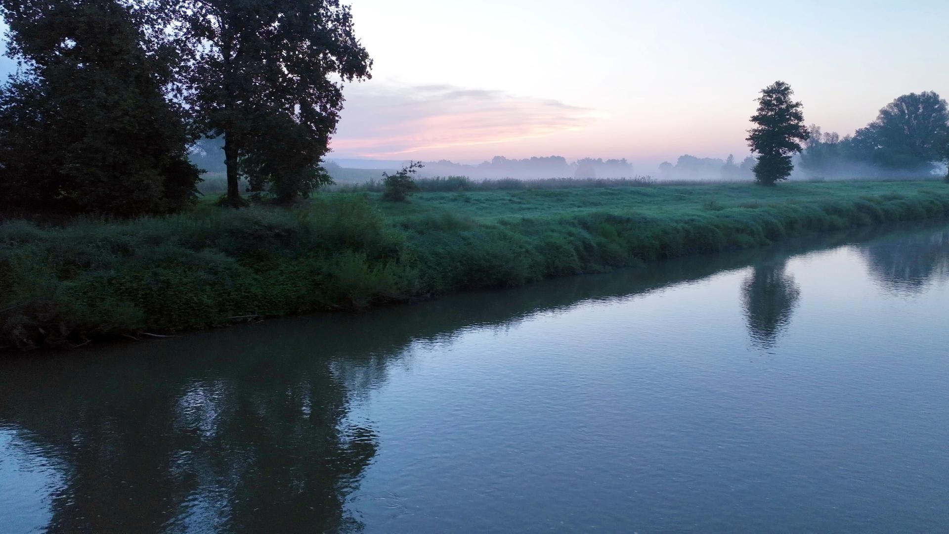 Ein Flusslauf mit Wiese und Bäumen im Morgennebel bei Sonnenaufgang