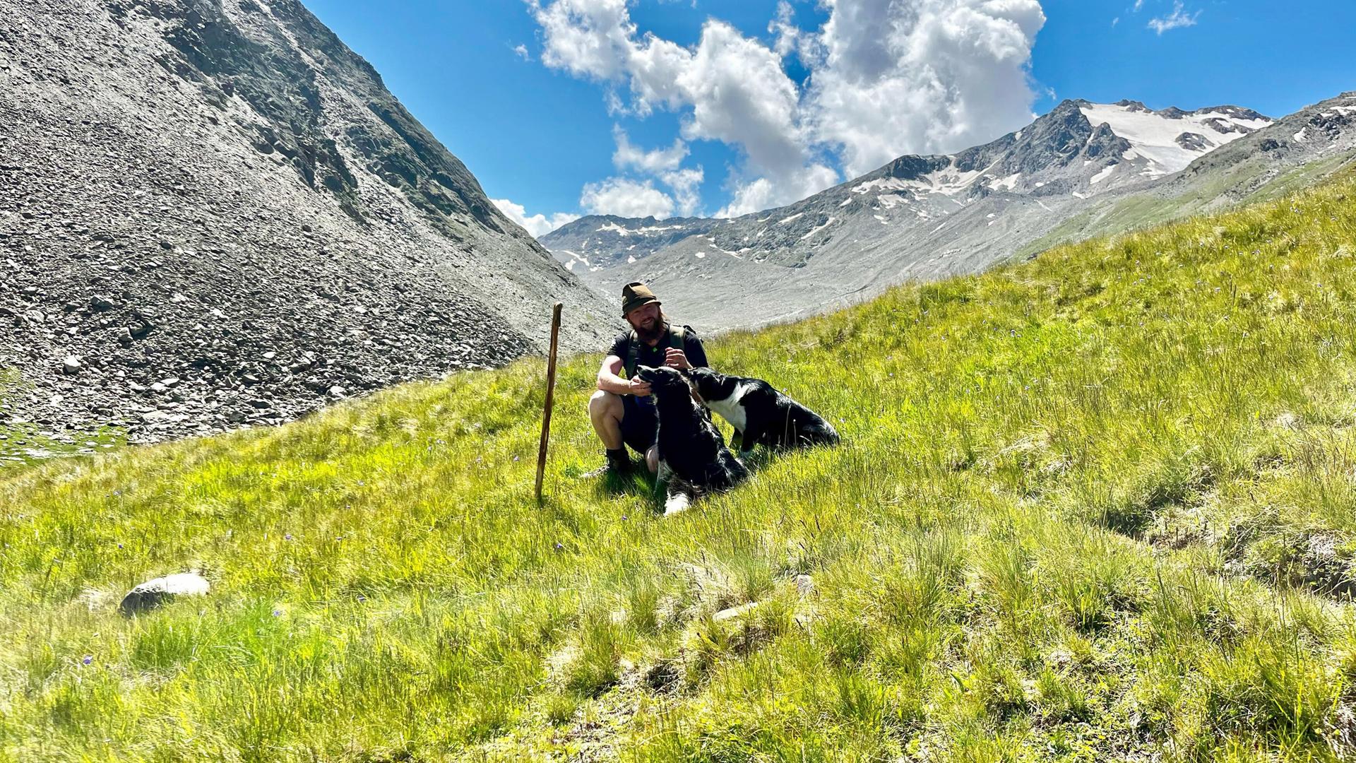 Ein Hirte sitzt mit seinen zwei Hunden auf einer grünen Bergwiese im Ötztal, im Hintergrund schneebedeckte Gipfel und blauer Himmel mit Wolken.