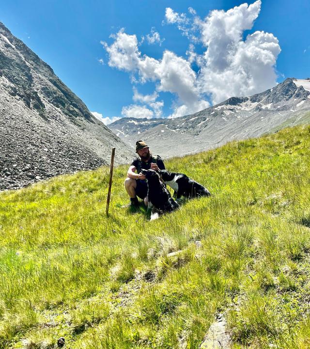 Ein Hirte sitzt mit seinen zwei Hunden auf einer grünen Bergwiese im Ötztal, im Hintergrund schneebedeckte Gipfel und blauer Himmel mit Wolken.