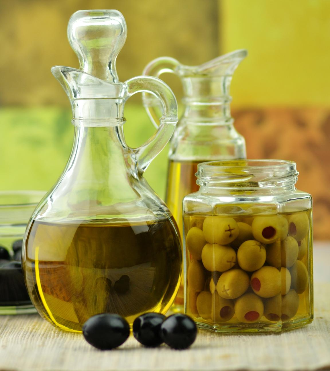 Olivenöl in einer Karaffe steht in der Mitte. Daneben steht ein Einmachglas mit Oliven und weitere Glasgefäße.