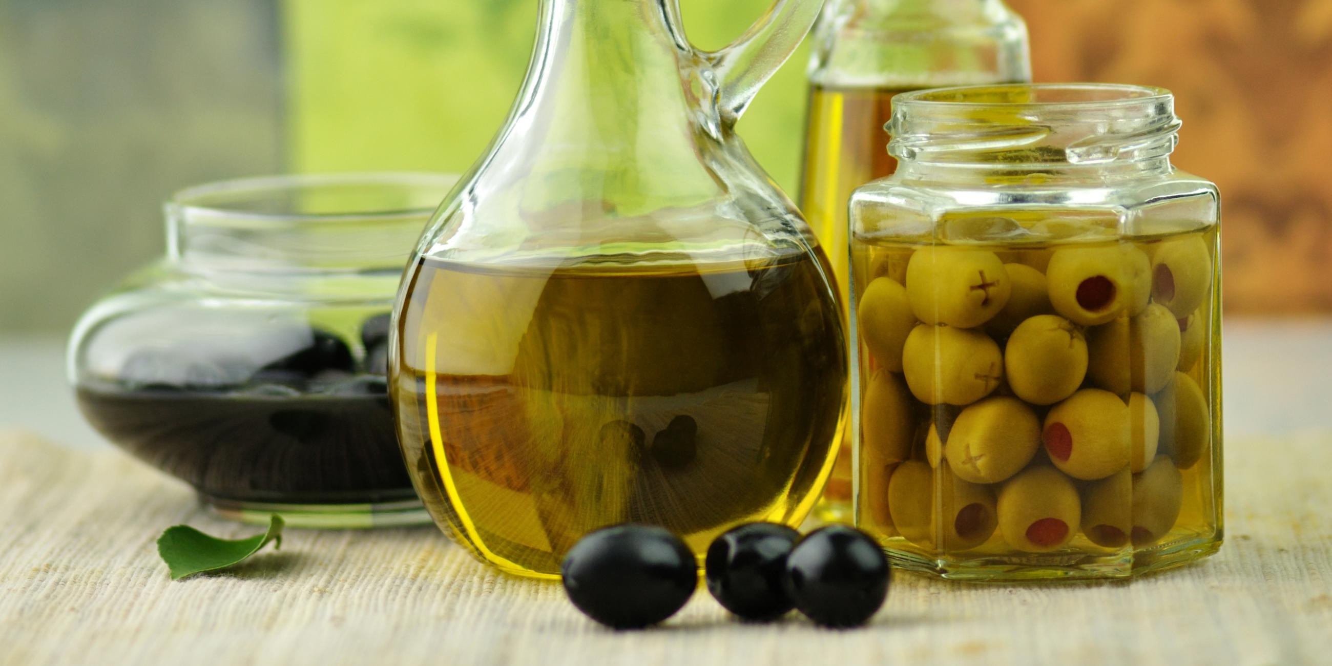 Olivenöl in einer Karaffe steht in der Mitte. Daneben steht ein Einmachglas mit Oliven und weitere Glasgefäße.