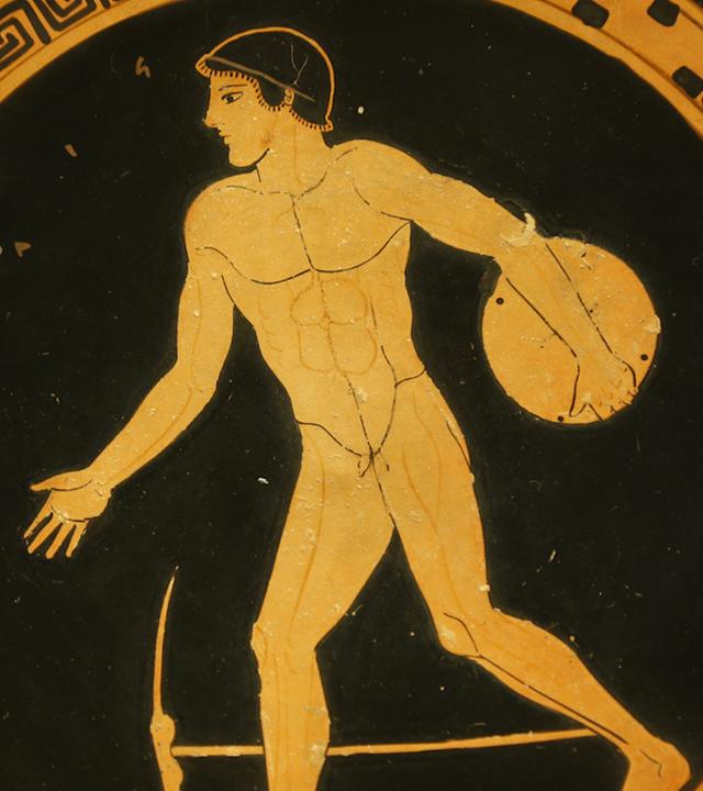 "Olympia: Die Wiege der Spiele": Farbbild einer griechischen Trinkschale. Ein nackter junger Mann in Schrittstellung nach links, der frontal wiedergegebene Oberkörper deutet die Drehung an. Der Sportler holt mit dem Diskus in seiner linken Hand hinter sich aus, die rechte ist nach vorn gestreckt. Zu seinen Füßen eine Hacke zum Aufrauhen oder Glätten des Bodens.