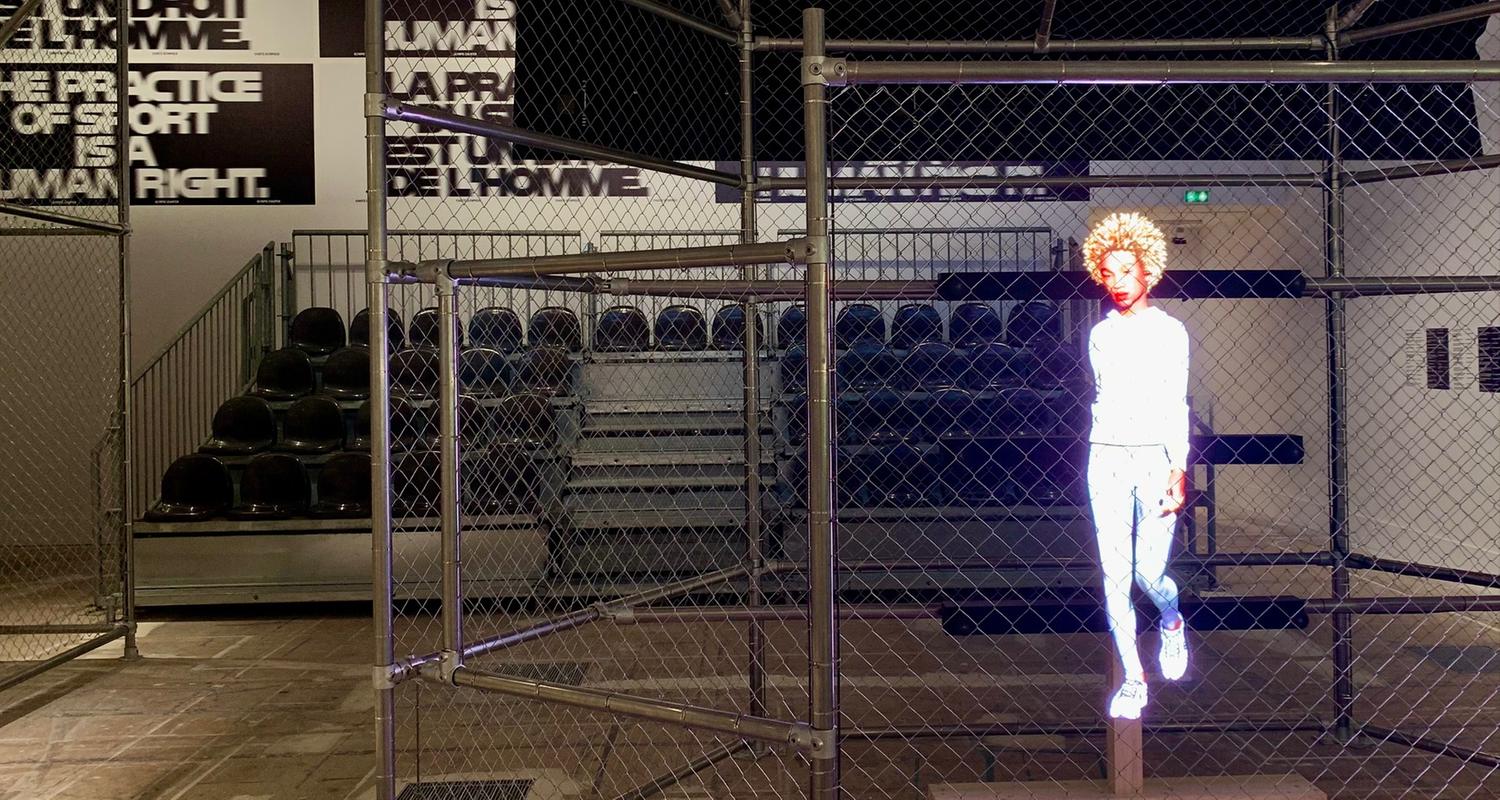 In einer Sporthalle sind Tribüne und Spielfeld mit Metallzäunen voneinander getrennt. Auf die Gitter wird ein Hologramm einer weiß gekleideten, sportlichen Frau projiziert.