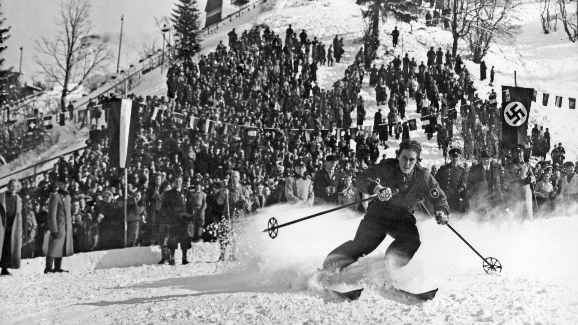 Historisches Schwarz-Weiß-Foto von einer Ski-Langläuferin, die vor einer Zuschauermenge den Berg hinunter saust. Am rechten Rand des Bildes ist eine Hakenkreuz-Flagge zu sehen.