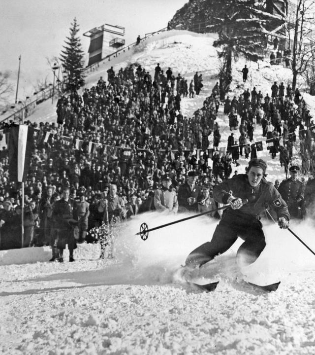Historisches Schwarz-Weiß-Foto von einer Ski-Langläuferin, die vor einer Zuschauermenge den Berg hinunter saust. Am rechten Rand des Bildes ist eine Hakenkreuz-Flagge zu sehen.