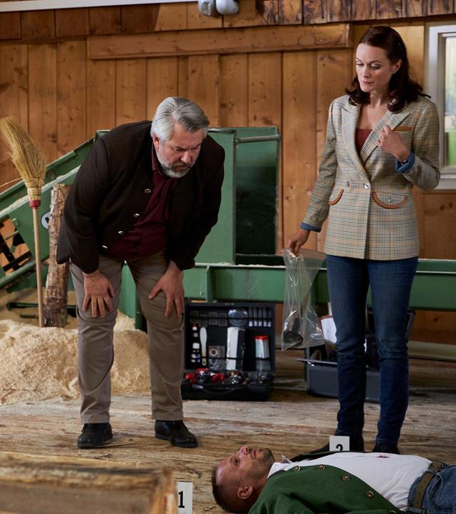 "Die Rosenheim-Cops - One Way in den Tod": Kommissarin Winter (Vanessa Eckart), Polizeihauptmeister Mohr (Max Müller), Kommissar Stadler (Dieter Fischer) und Pathologin Mai (Sina Wilke) stehen in einem Holzhaus. Vor ihnen liegt das Mordopfer auf dem Rücken. Im Hintergrund ist ein Haufen Sägespäne sowie eine dunkelgrüne Maschine zu sehen.