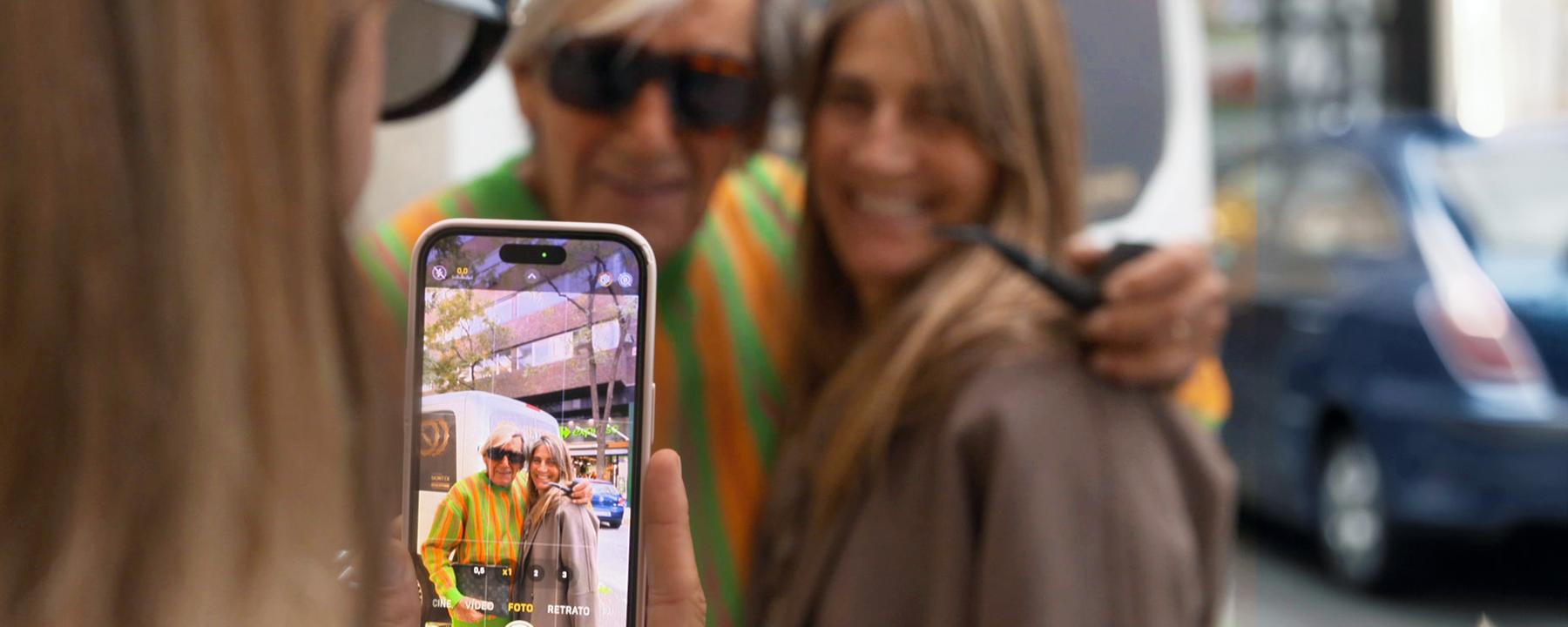 Fotosession mit einem Handy. Der leuchtende Handy-Bildschirm zeigt einen, älteren Mann mit Sonnenbrille, neben ihm eine jüngere Frau mit langen Haaren. Im Hintergrund die beiden, die gerade fotografiert werden, noch einmal unscharf zu sehen.