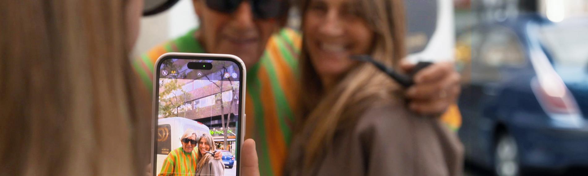 Fotosession mit einem Handy. Der leuchtende Handy-Bildschirm zeigt einen, älteren Mann mit Sonnenbrille, neben ihm eine jüngere Frau mit langen Haaren. Im Hintergrund die beiden, die gerade fotografiert werden, noch einmal unscharf zu sehen.