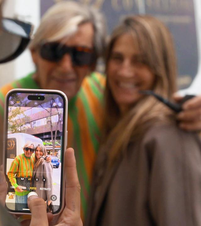 Fotosession mit einem Handy. Der leuchtende Handy-Bildschirm zeigt einen, älteren Mann mit Sonnenbrille, neben ihm eine jüngere Frau mit langen Haaren. Im Hintergrund die beiden, die gerade fotografiert werden, noch einmal unscharf zu sehen.