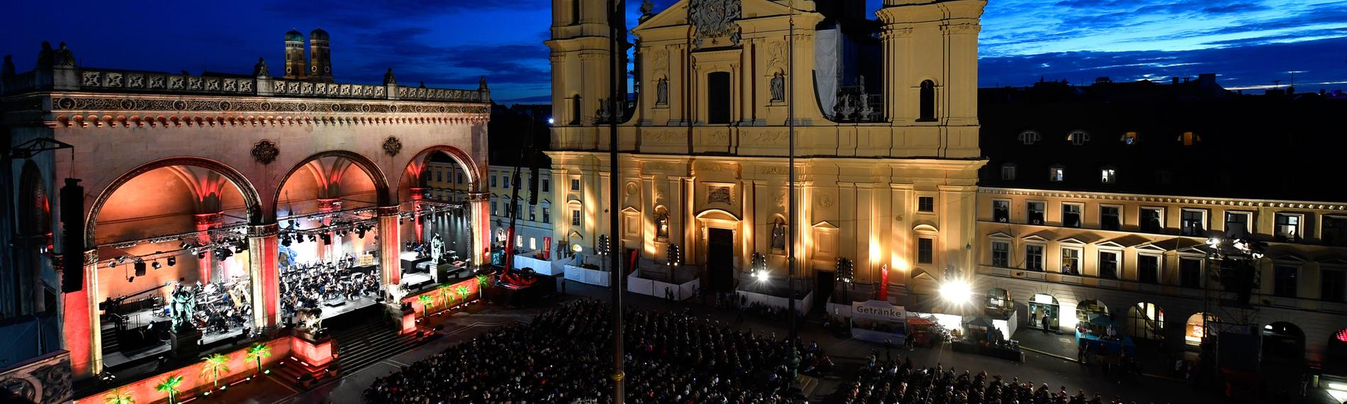 Der Münchner Odeonsplatz in der Abenddämmerung mit sitzendem Publikum. Theatinerkirche und Feldherrnhalle sind beleuchtet. In der Feldherrnhalle sitzen die Münchner Philharmoniker.