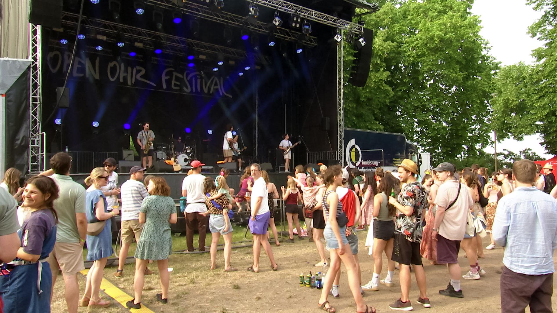 Vor einer Festivalbühne, auf der Musiker spielen, stehen Menschen.