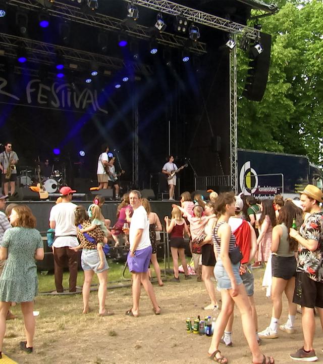 Vor einer Festivalbühne, auf der Musiker spielen, stehen Menschen.