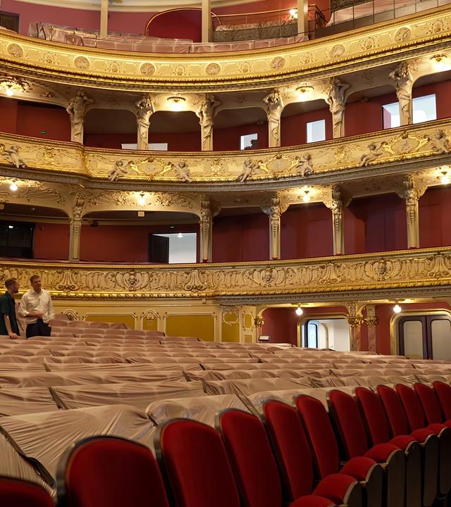 Das Bild zeigt den Innenraum eines Theaters. Im Vordergrund sind Sitzreihen aus gepolstertem Stoff zu sehen, die teilweise mit transparentem Plastik abgedeckt sind. Zwei Männer stehen im Bild; einer von ihnen hat einen kurzen Bart und trägt ein dunkelgrünes Hemd, der andere trägt ein weißes Hemd und hat kurz geschnittene Haare. Der Mann im weißen Hemd zeigt mit der Hand in eine Richtung, während der andere ihn aufmerksam anschaut.   Die Wände des Theaters sind reich verziert mit goldenen Ornamenten und Skulpturen. Die Balustrade, die die Sitzreihen umfasst, hebt sich durch ihren filigranen Kunststil hervor. Deckenlichter sind sichtbar, und der Raum wirkt gut beleuchtet, während im Hintergrund große Fenster zu erkennen sind. Es handelt sich um einen eleganten und historischen Raum, der für Aufführungen konzipiert ist.