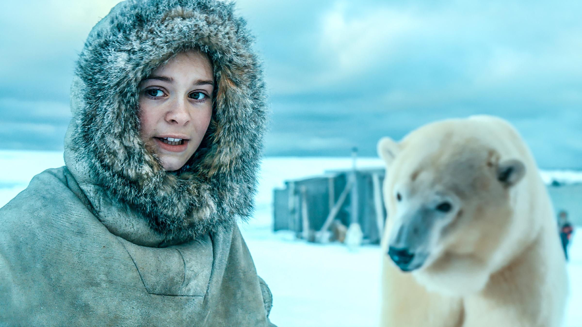 Links das Mädchen Julia (Kaisa Gurine Antonsen) mit einer Fell umrandeten Kapuze im Schnee, rechts ein Eisbär, der ihr schon ziemlich nahe kommt. Im Hintergrund ist eine Hütte zu sehen.