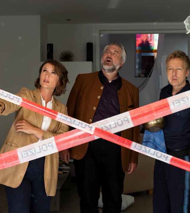 Die Kommissare Andresen (Sophie Melbinger) und Stadler (Dieter Fischer) stehen mit Polizeihauptmeister Mohr (Max Müller) in einem Wohnraum und begutachten ein zerbrochenes Fenster, das mit rot-weißem Flatterband der Polizei abgesperrt ist.