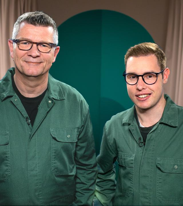 Zwei Menschen in grünen Overalls blicken in die Kamera.