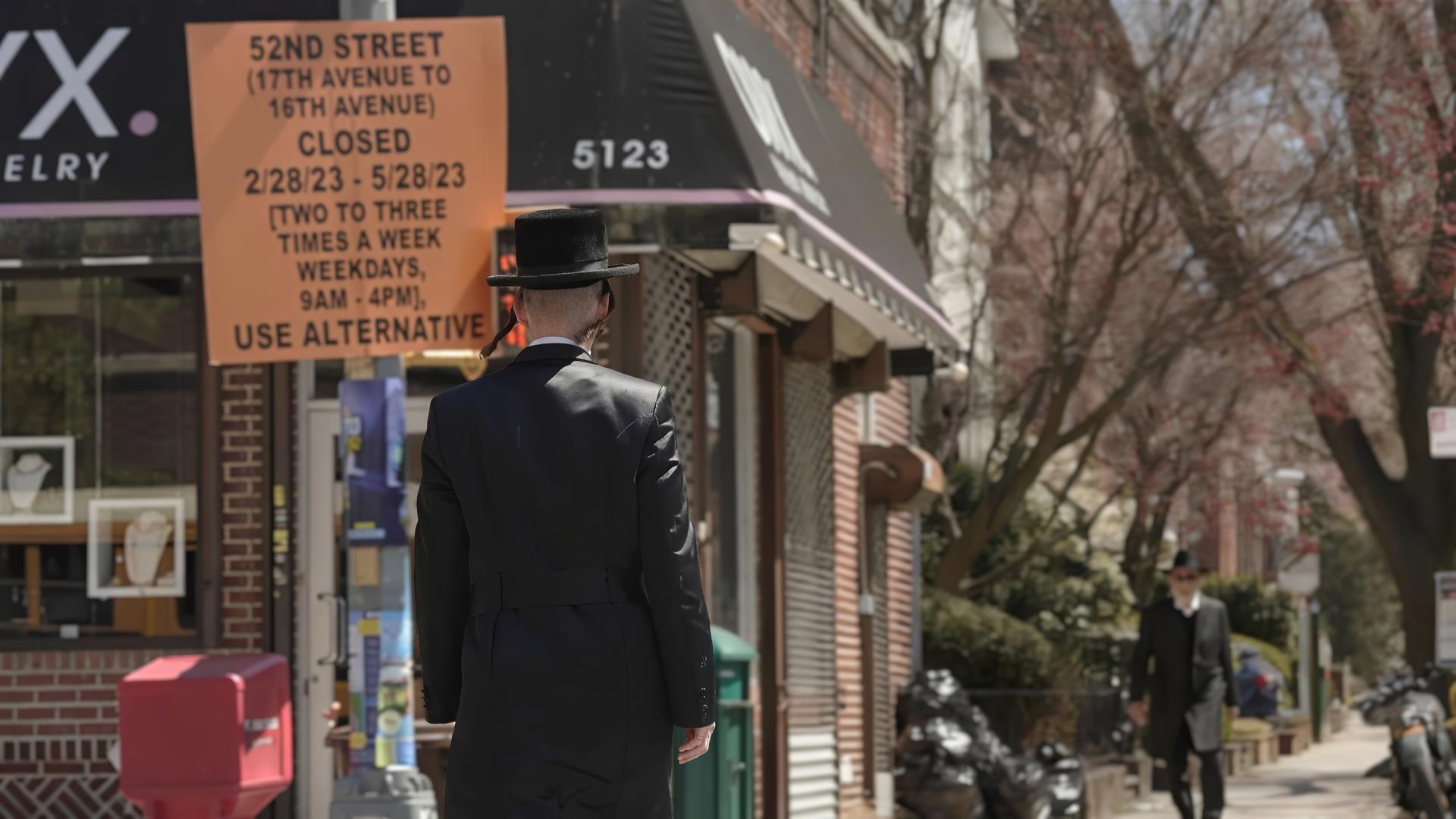 Mann in schwarzer Anzugjacke, mit Schläfenlocken und schwarzem Hut läuft auf Straße an einem Mast vorbei, daran hängt ein orangenfarbenes Schild mit schwarzer Aufschrift (Ankündigung von Straßensperrung); dahinter ein Laden mit schwarzer Markise, kahle Bäume im Hintergrund. 