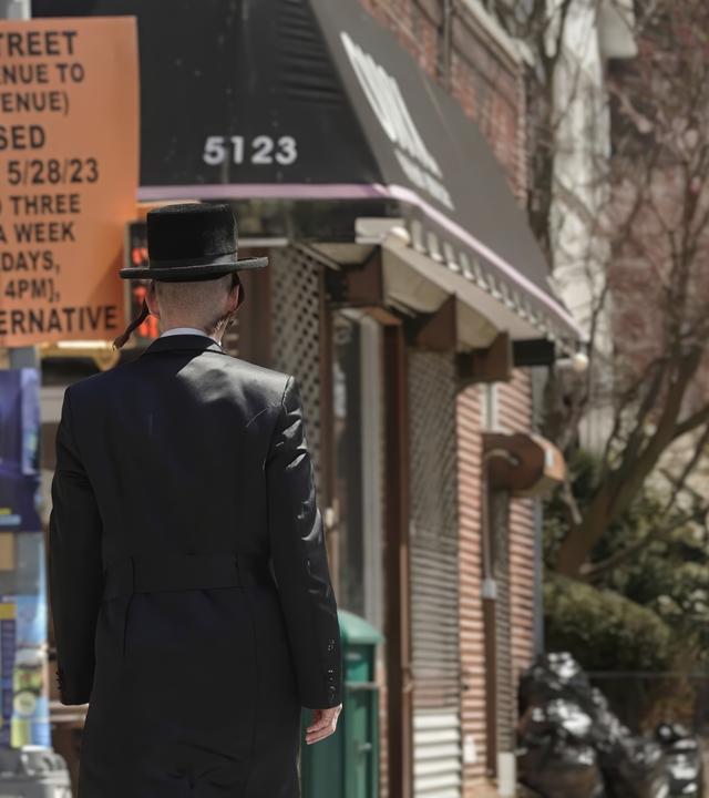 Mann in schwarzer Anzugjacke, mit Schläfenlocken und schwarzem Hut läuft auf Straße an einem Mast vorbei, daran hängt ein orangenfarbenes Schild mit schwarzer Aufschrift (Ankündigung von Straßensperrung); dahinter ein Laden mit schwarzer Markise, kahle Bäume im Hintergrund. 