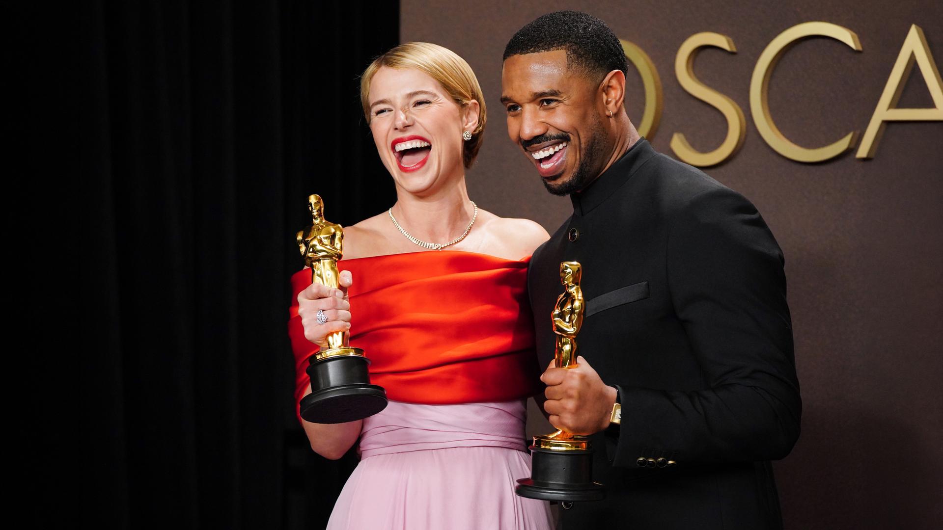 Eine Frau im Anebdkleid und ein Mann in Smoking halten goldene Preise in Form einer Männerstatue in den Händen und lachen.