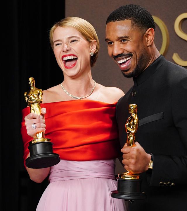 Eine Frau im Anebdkleid und ein Mann in Smoking halten goldene Preise in Form einer Männerstatue in den Händen und lachen.