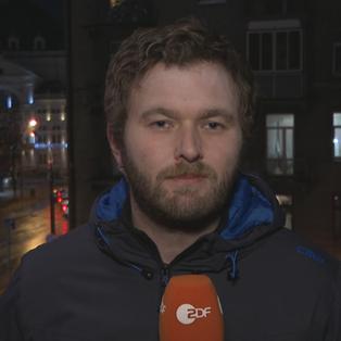 ZDF-Korrepondent Sebastian Ehm vor städtischer Kulise mit Mikrofon mit einem ZDF-Logo in der Hand