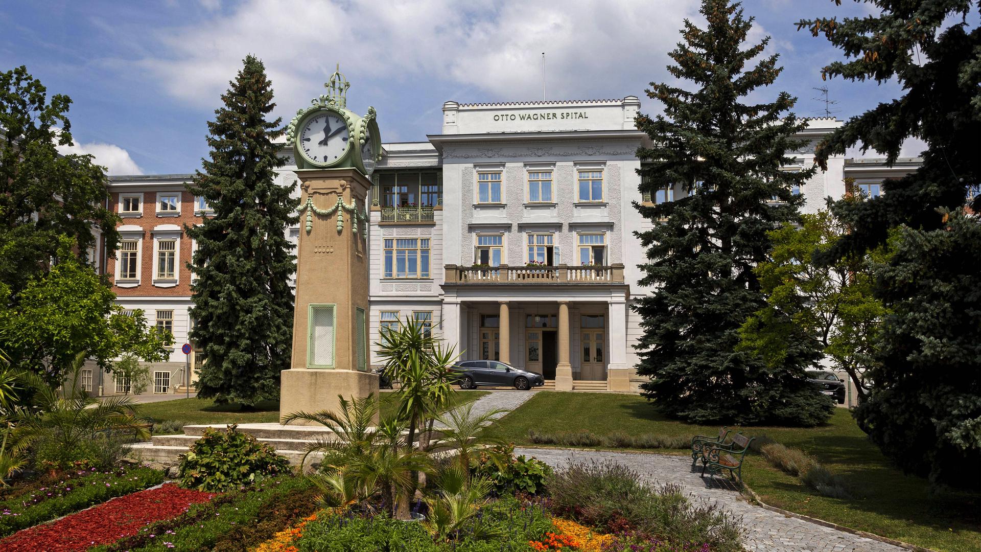 Ein großes altes Gebäude mit Säuleneingang. Davor Blumenrabatten und eine Uhr auf einer hohen Säule.