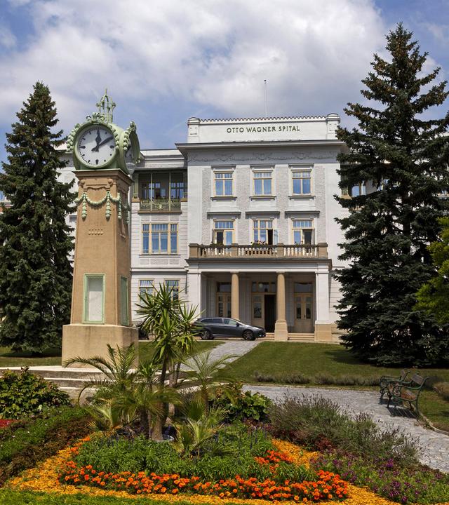 Ein großes altes Gebäude mit Säuleneingang. Davor Blumenrabatten und eine Uhr auf einer hohen Säule.