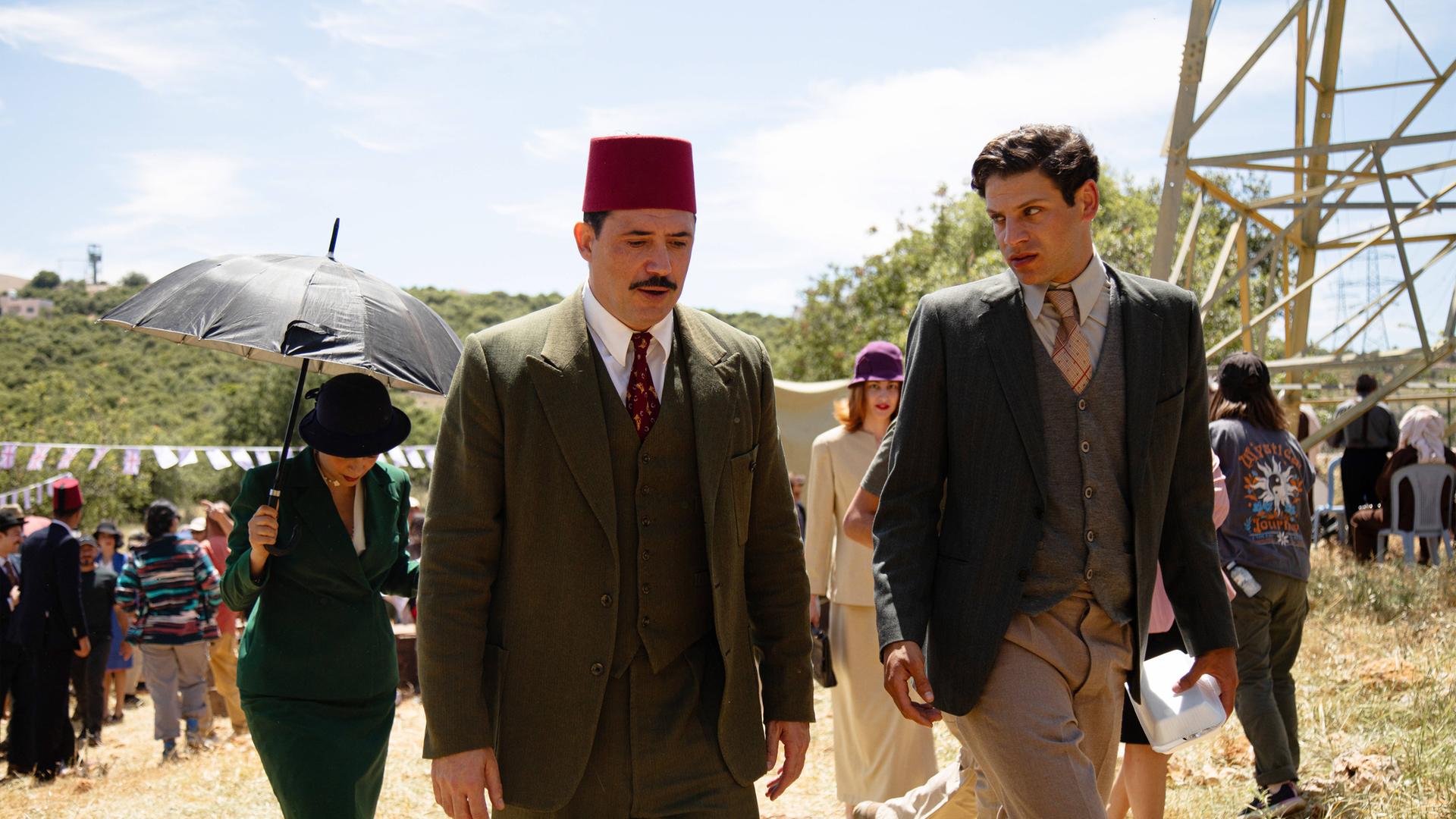 Zwei Männer laufen nebeneinander her und unterhalten sich. Beide tragen Anzüge, einer hat einen Fez als Kopfbedeckung. Im Hintergrund sind andere Menschen zu sehen.