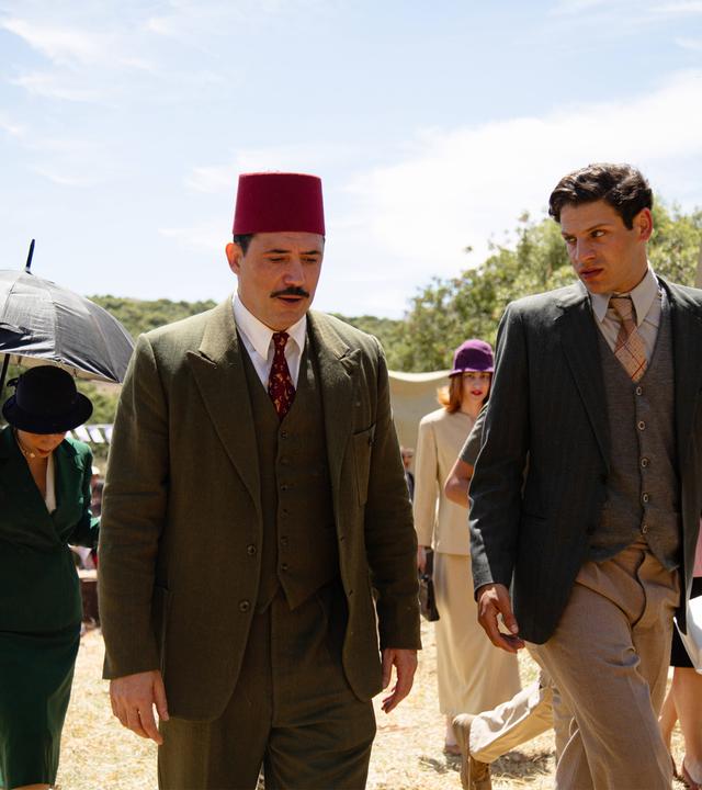 Zwei Männer laufen nebeneinander her und unterhalten sich. Beide tragen Anzüge, einer hat einen Fez als Kopfbedeckung. Im Hintergrund sind andere Menschen zu sehen.