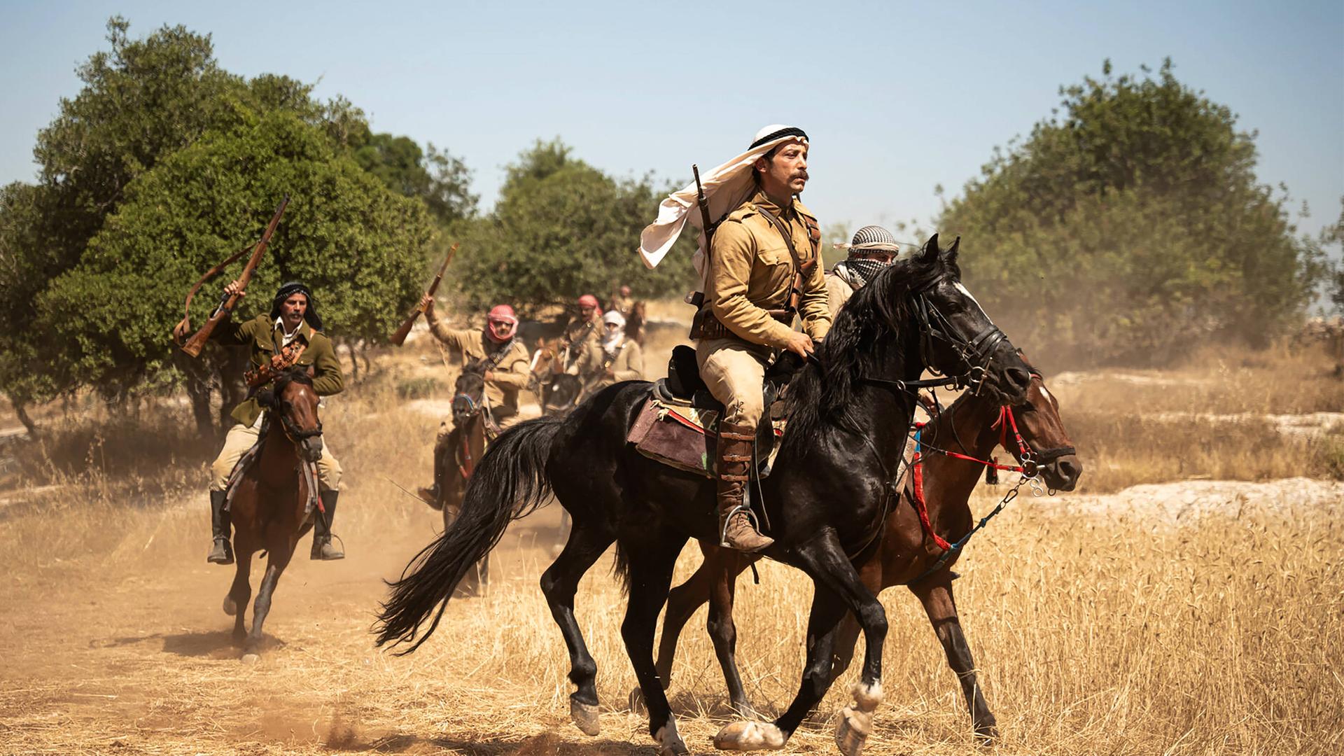 Männer mit erhobenen Gewehren reiten auf Pferden durch eine trockene Landschaft.