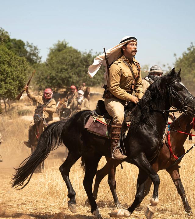 Männer mit erhobenen Gewehren reiten auf Pferden durch eine trockene Landschaft.