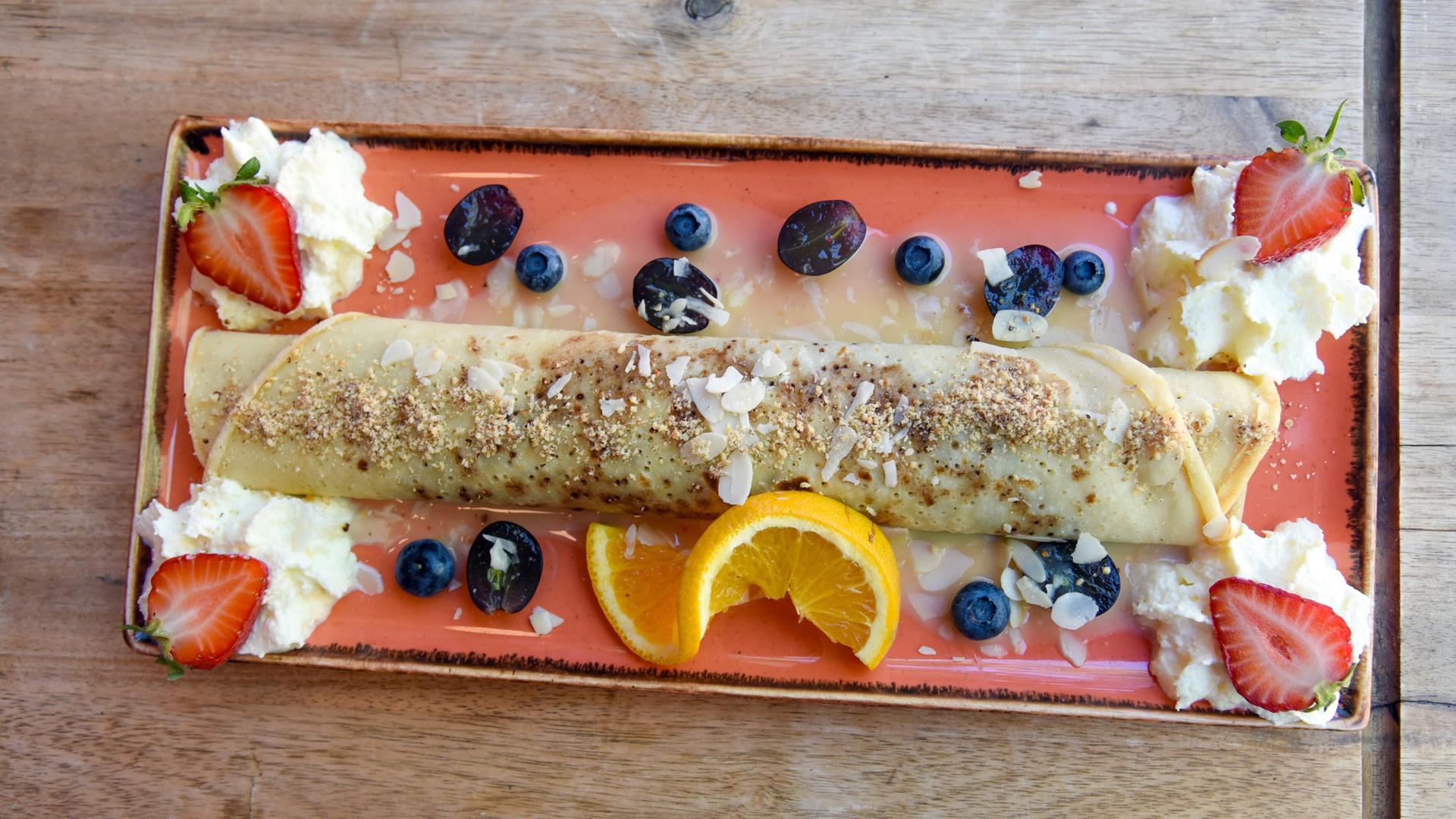 Das Bild zeigt einen Teller mit einer gefüllten Palatschinke, die auf einem farbigen, rechteckigen Teller präsentiert ist. Die Palatschinke ist rötlich-braun und aufgerollt, mit einer Mischung aus gehackten, gerösteten Nüssen und mandelartigen Streuseln bestreut. Um die Palatschinke herum befinden sich mehrere frische Früchte: zwei Erdbeeren, einige Heidelbeeren und eine halbe orange Scheibe. Auf einer Seite des Tellers gibt es einen Klecks Sahne, der in zwei Portionen verteilt ist. Die Unterlage des Tellers zeigt eine Holzoberfläche, die zur rustikalen Präsentation beiträgt.